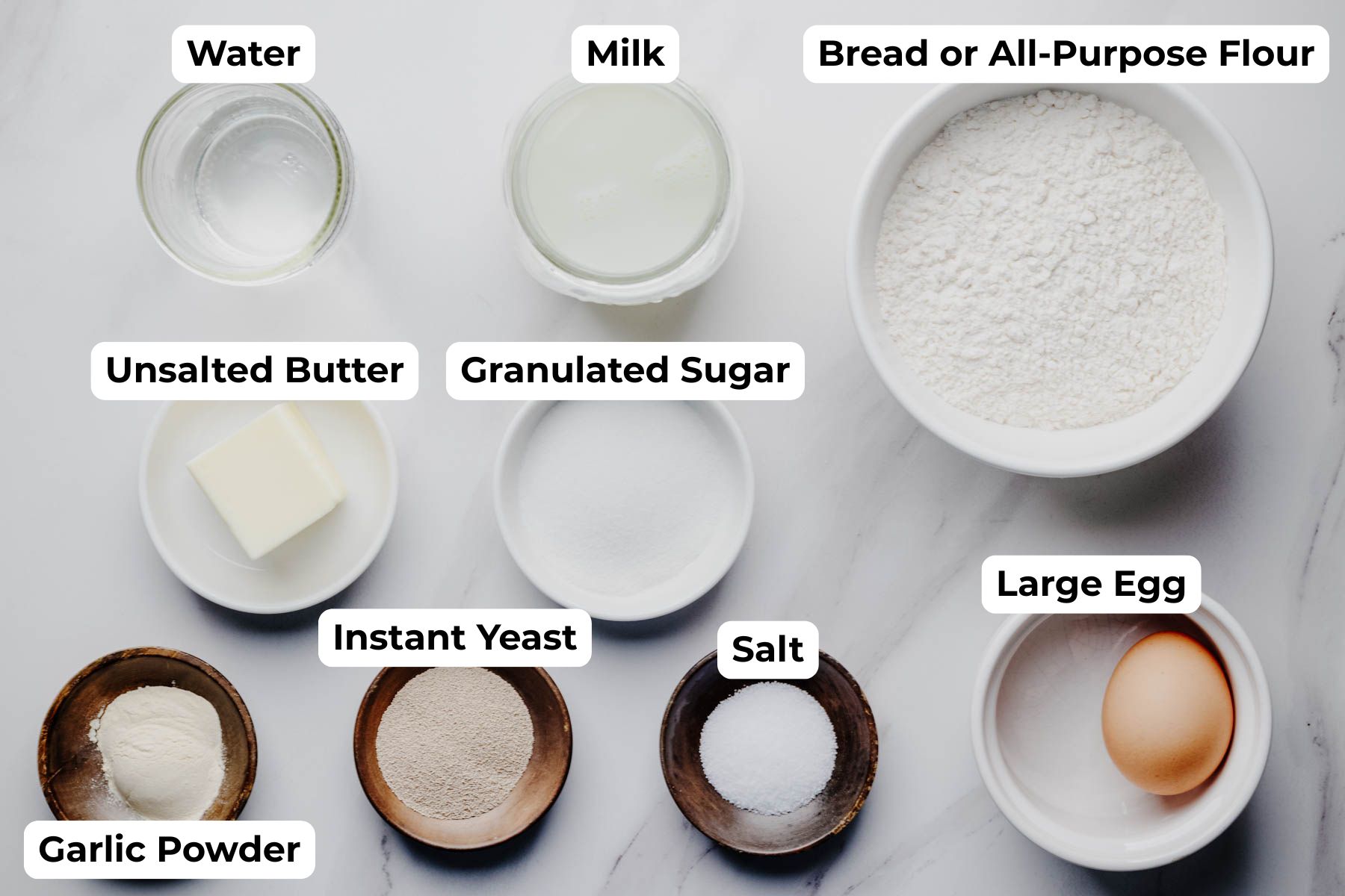 Overhead view of the labeled ingredients for garlic dinner rolls, including water, milk, flour, butter, sugar, garlic powder, instant yeast, salt, and one large egg.