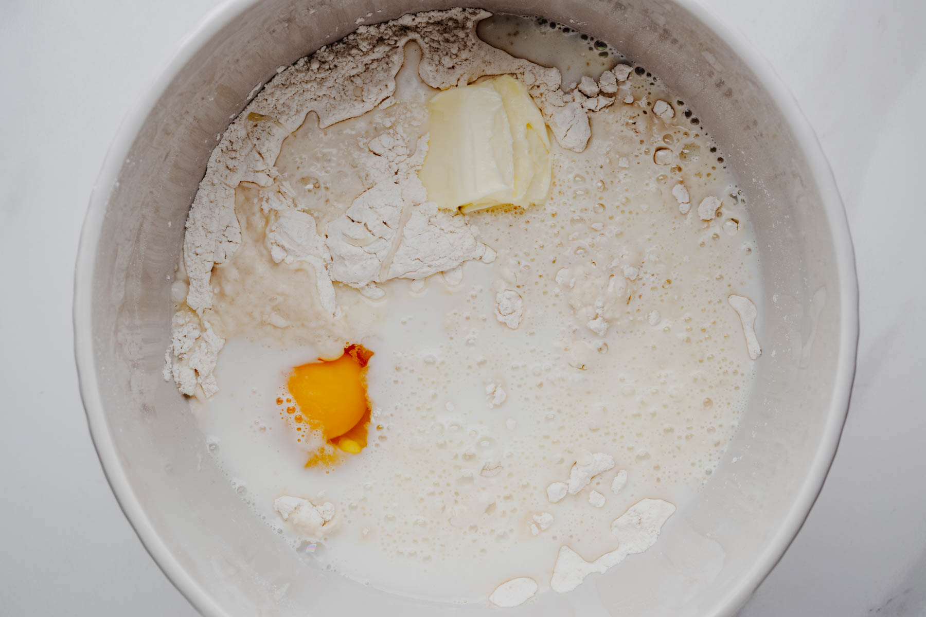 Overhead view of warm milk, water, butter, and egg added to the dry ingredients for garlic dinner roll dough.