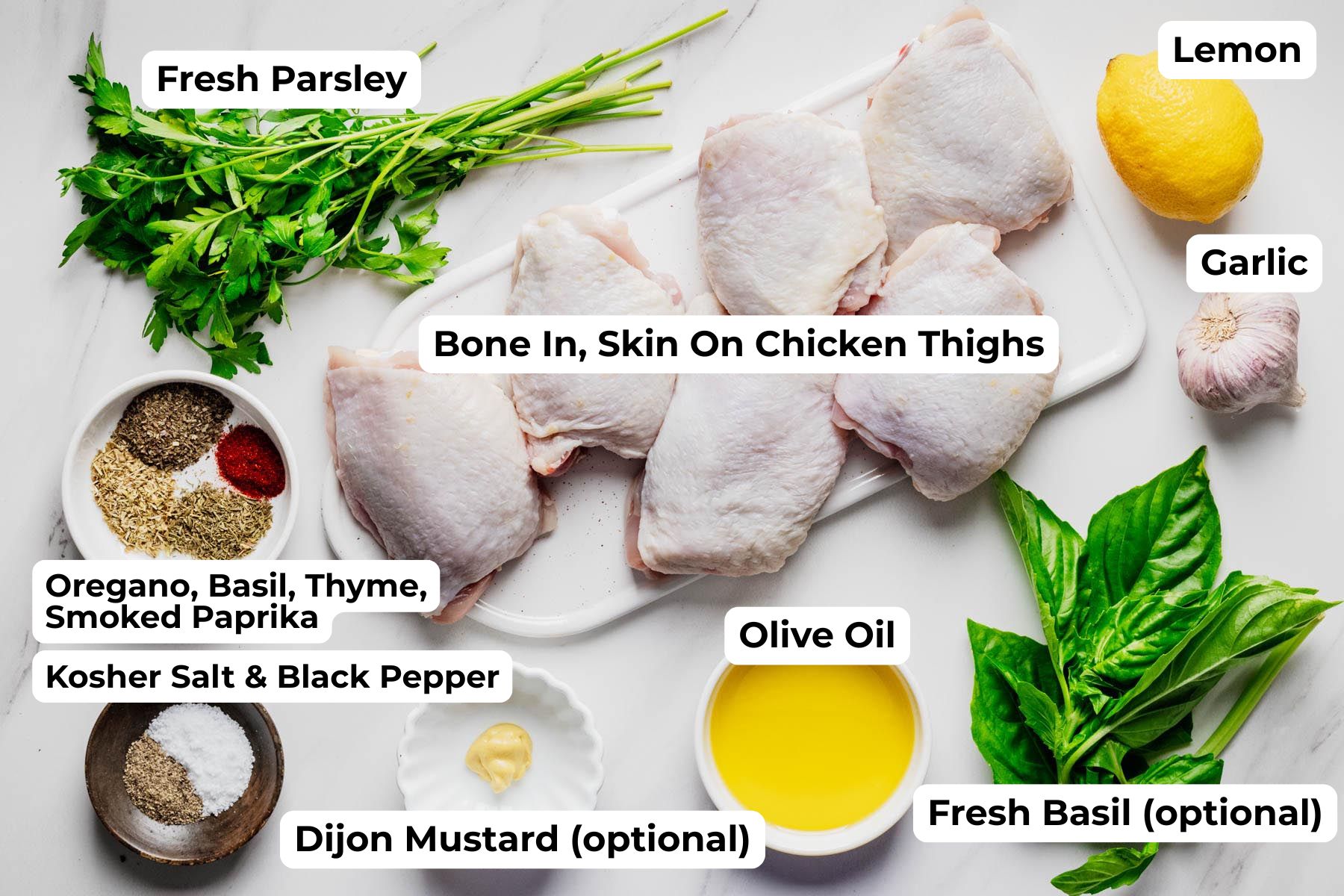 Labeled ingredient photo showing bone-in, skin-on chicken thighs, fresh parsley, lemon, garlic, olive oil, fresh basil, Dijon mustard, kosher salt, black pepper, oregano, basil, thyme, and smoked paprika arranged on a white surface for grilled lemon herb chicken thighs.