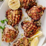 Grilled lemon herb chicken thighs arranged on a white serving platter with lemon wedges, fresh parsley, and charred lemon halves.
