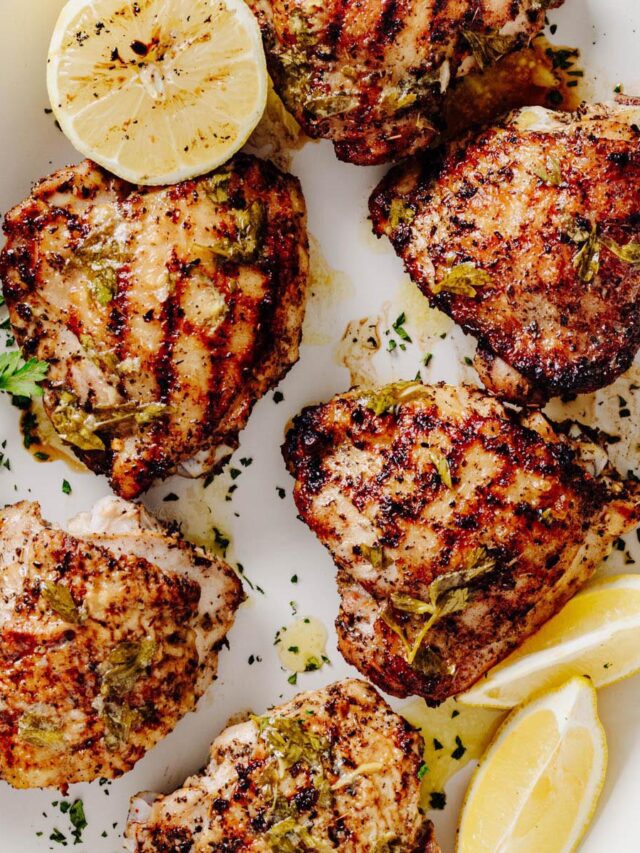 Grilled lemon herb chicken thighs arranged on a white serving platter with lemon wedges, fresh parsley, and charred lemon halves.