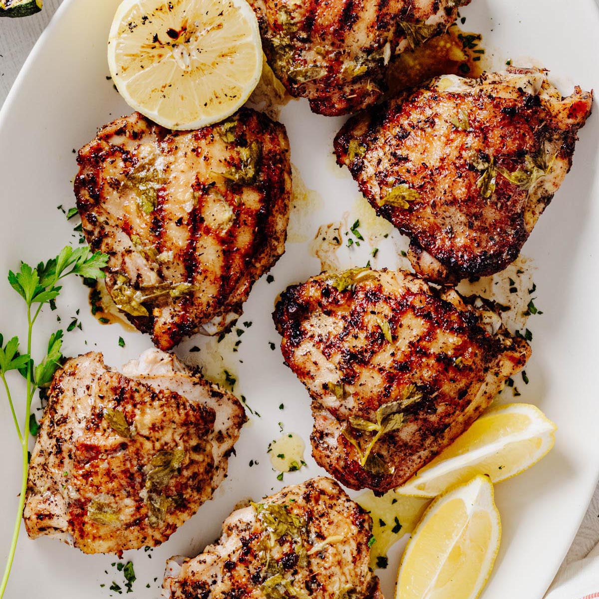 Grilled lemon herb chicken thighs arranged on a white serving platter with lemon wedges, fresh parsley, and charred lemon halves.
