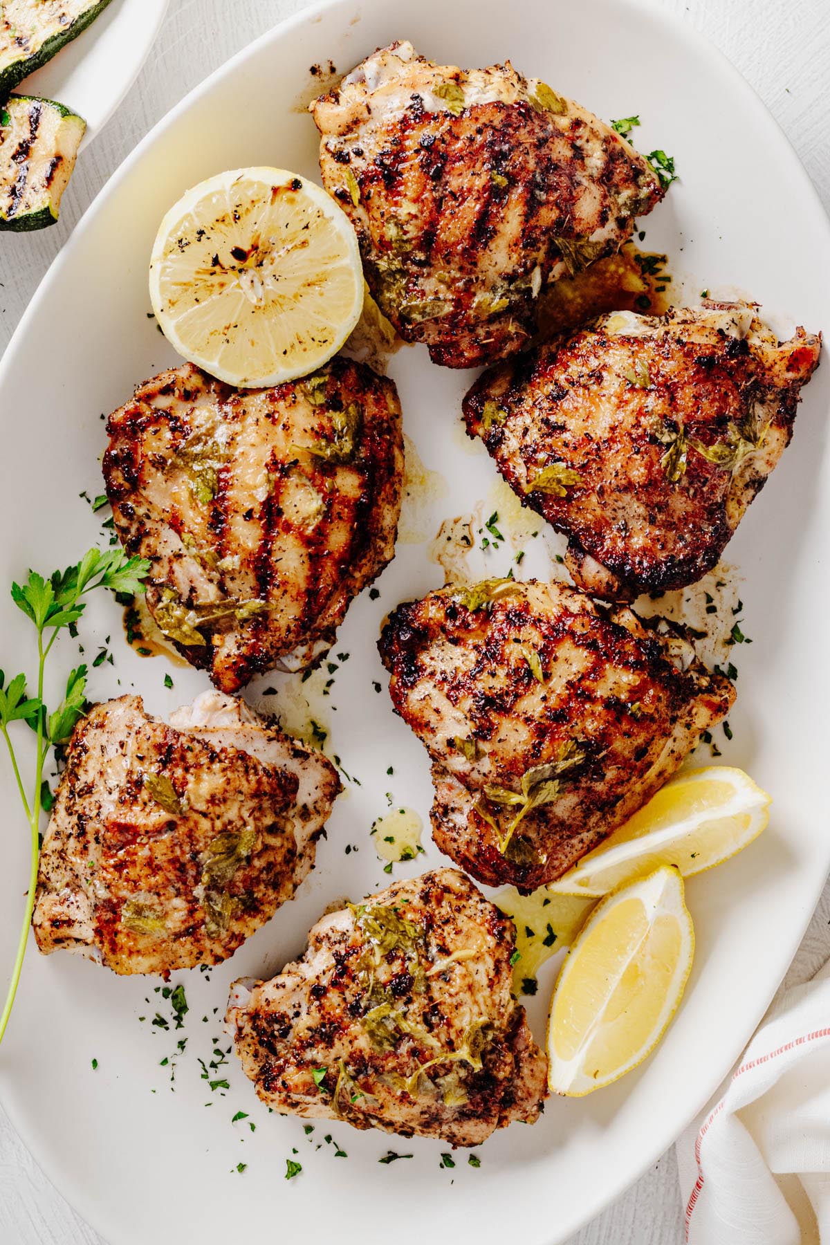 Grilled lemon herb chicken thighs arranged on a white serving platter with lemon wedges, fresh parsley, and charred lemon halves.