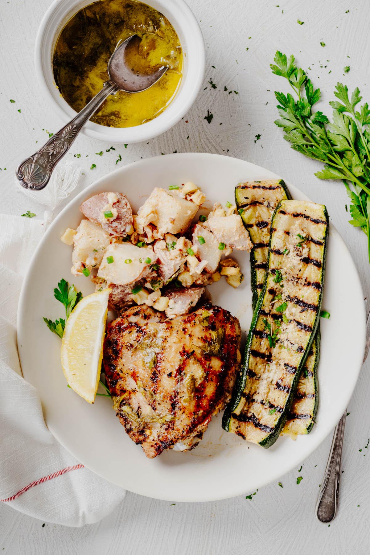 Plate of grilled lemon herb chicken thighs served with grilled Parmesan zucchini, creamy potato salad, a lemon wedge, and a bowl of lemon herb finishing sauce.