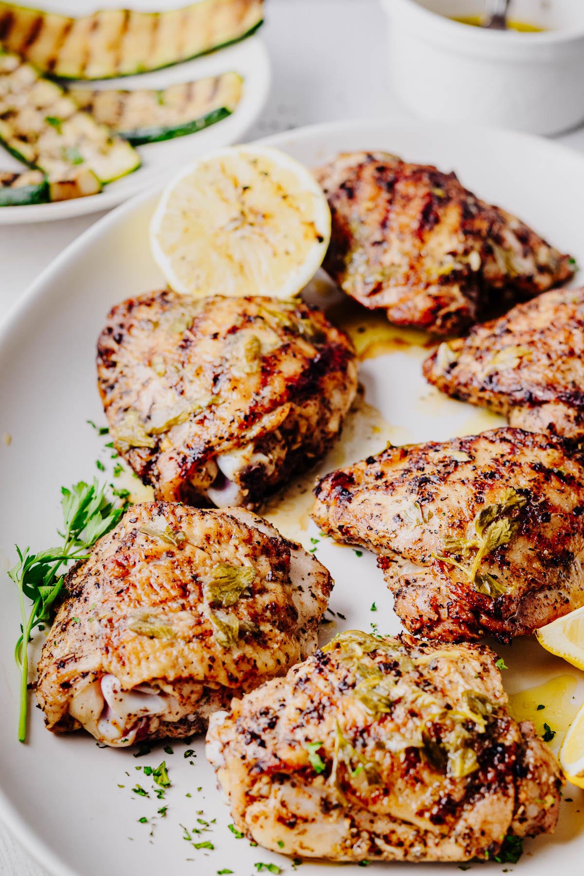 A platter of grilled lemon herb chicken thighs with crispy, golden-brown skin, fresh parsley, grilled lemon, and a small dish of Parmesan grilled zucchini blurred in the background.