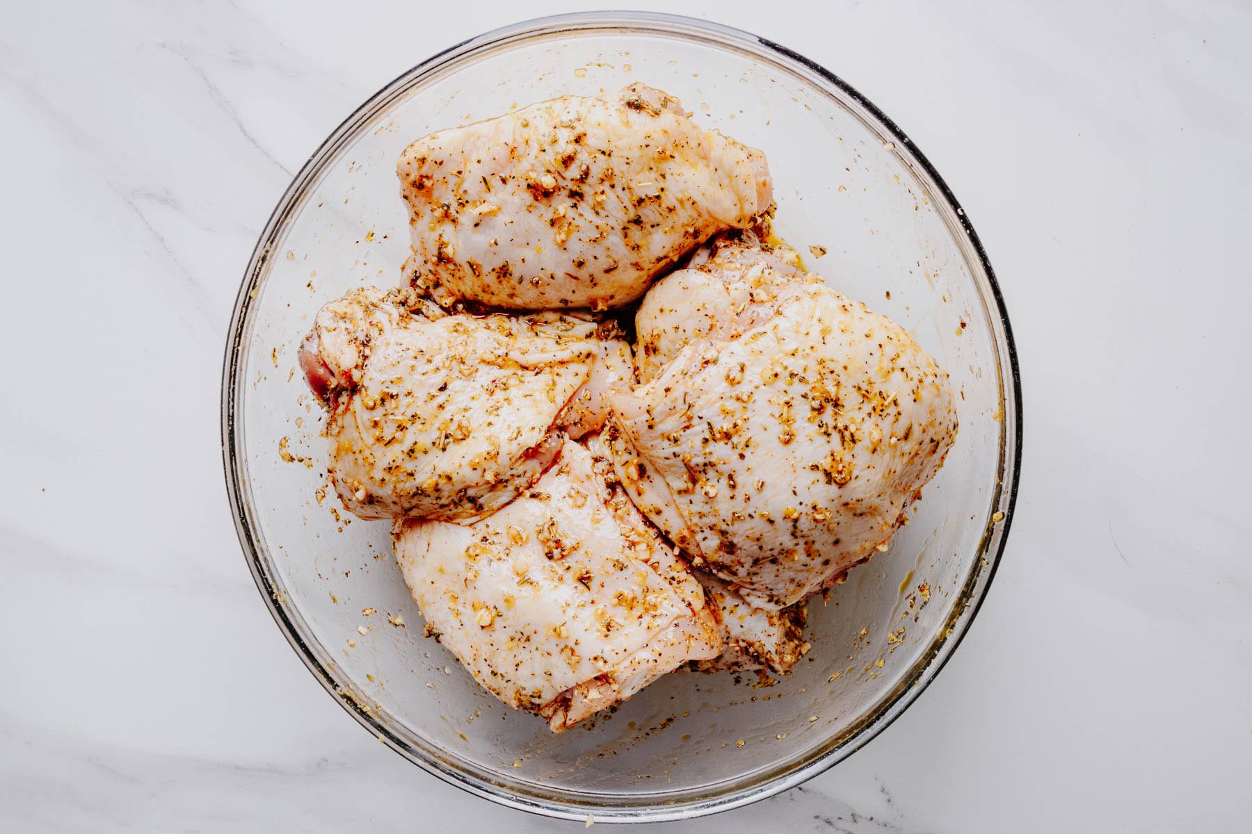Bone-in, skin-on chicken thighs coated in lemon herb seasoning in a glass mixing bowl before grilling.