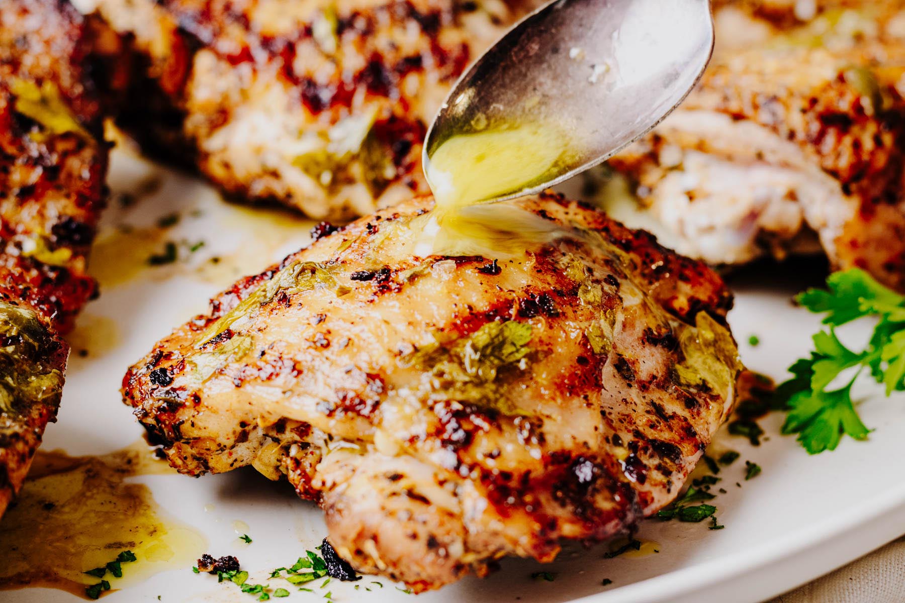 A spoon drizzling lemon herb finishing sauce over grilled chicken thighs on a serving platter.