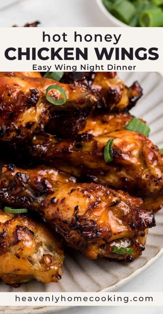 A plate of crispy hot honey chicken wings is coated in a glossy sweet and spicy glaze and garnished with sliced green onions. This vertical Pinterest pin features a homemade hot honey chicken wings recipe from Heavenly Home Cooking.