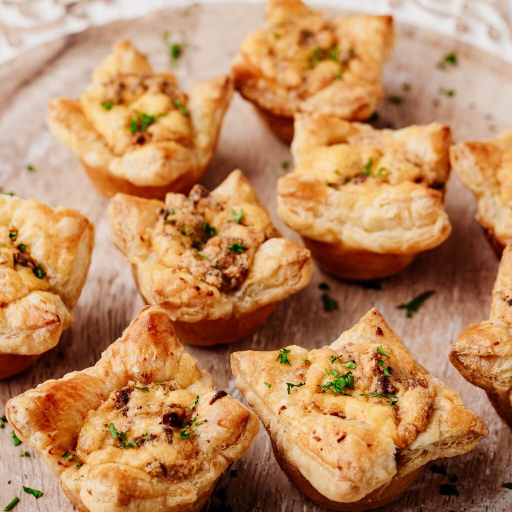 Sausage puff pastry cups filled with creamy Italian sausage and cheddar, garnished with chopped parsley on a wooden serving board.