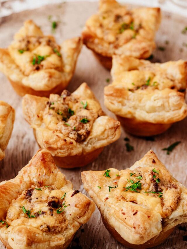 Sausage puff pastry cups filled with creamy Italian sausage and cheddar, garnished with chopped parsley on a wooden serving board.