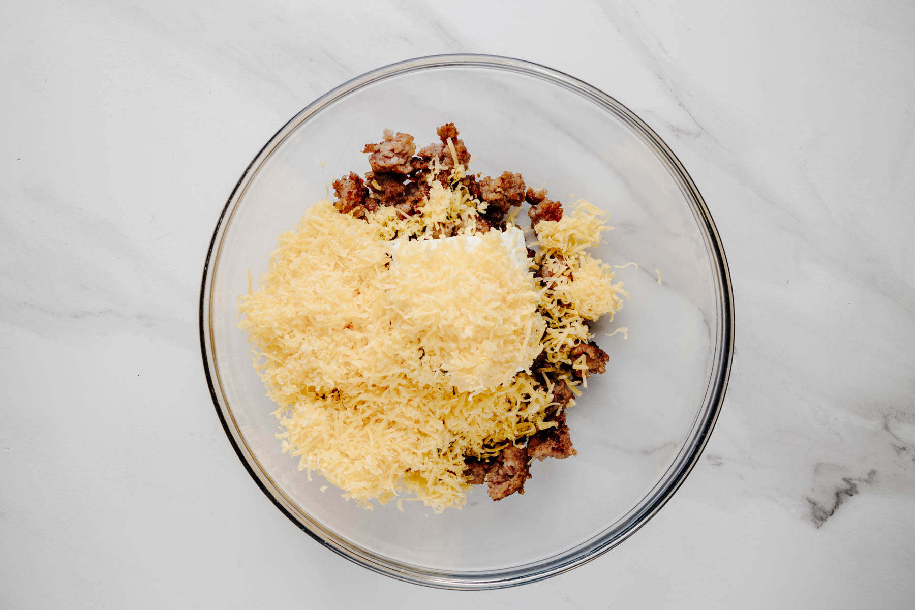 Cooked Italian sausage, softened cream cheese, shredded cheddar, garlic powder, and black pepper in a glass mixing bowl.