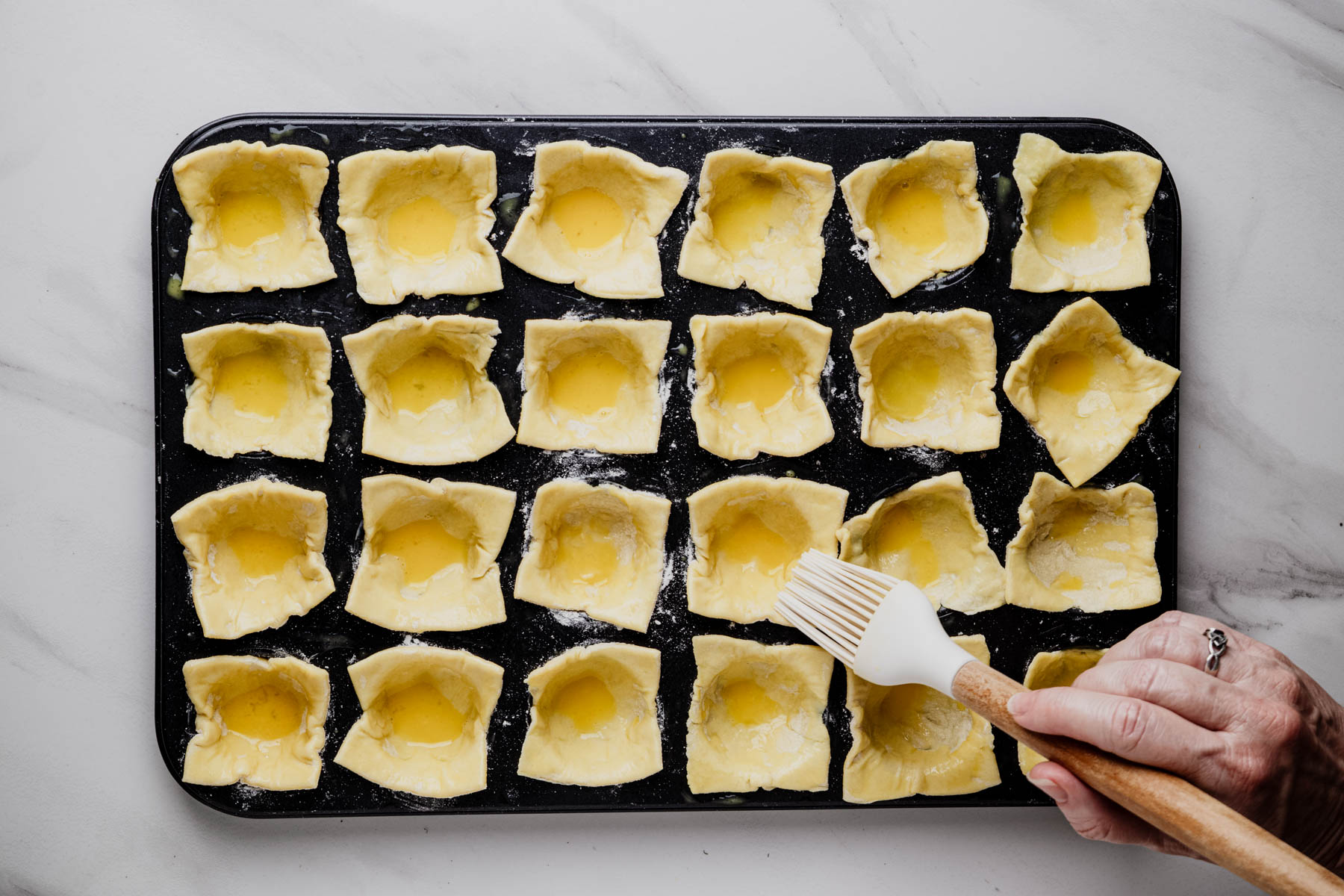 Egg wash being brushed over puff pastry squares in a mini muffin pan.