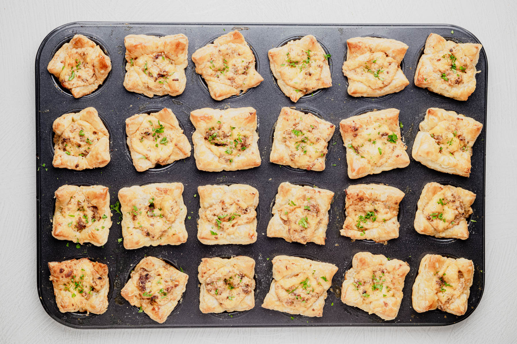 Golden sausage puff pastry cups garnished with chopped parsley in a mini muffin pan.