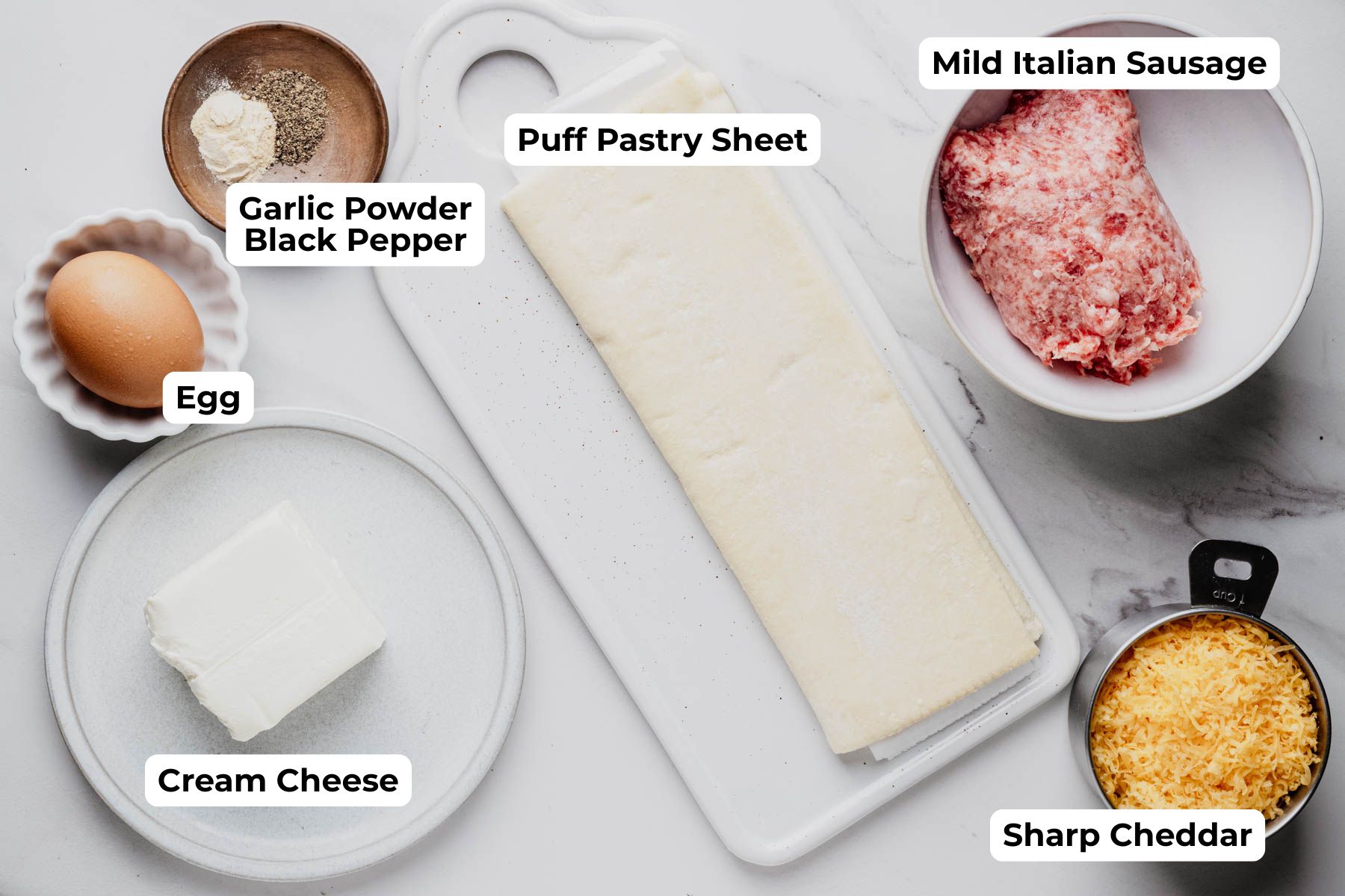 Overhead view of the ingredients for sausage puff pastry cups, including puff pastry, mild Italian sausage, cream cheese, sharp cheddar, an egg, garlic powder, and black pepper, with each ingredient labeled.