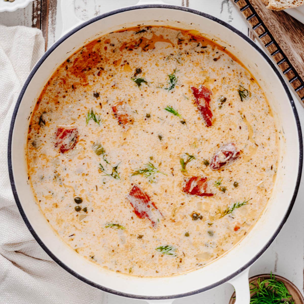 Overhead view of creamy smoked salmon chowder with salmon pieces, capers, and fresh dill in a white Dutch oven.