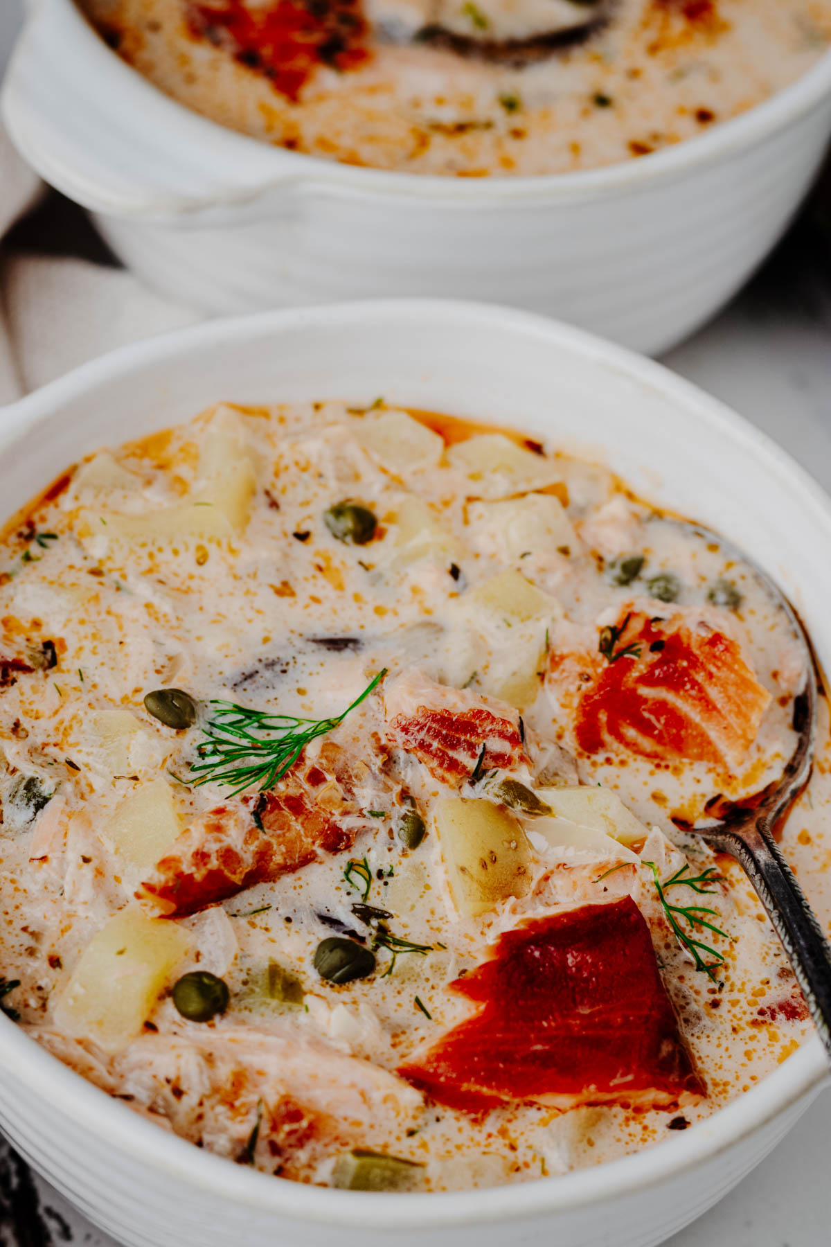 Close-up of chunky smoked salmon chowder with yellow potatoes, capers, fresh dill, and a spoonful of salmon.