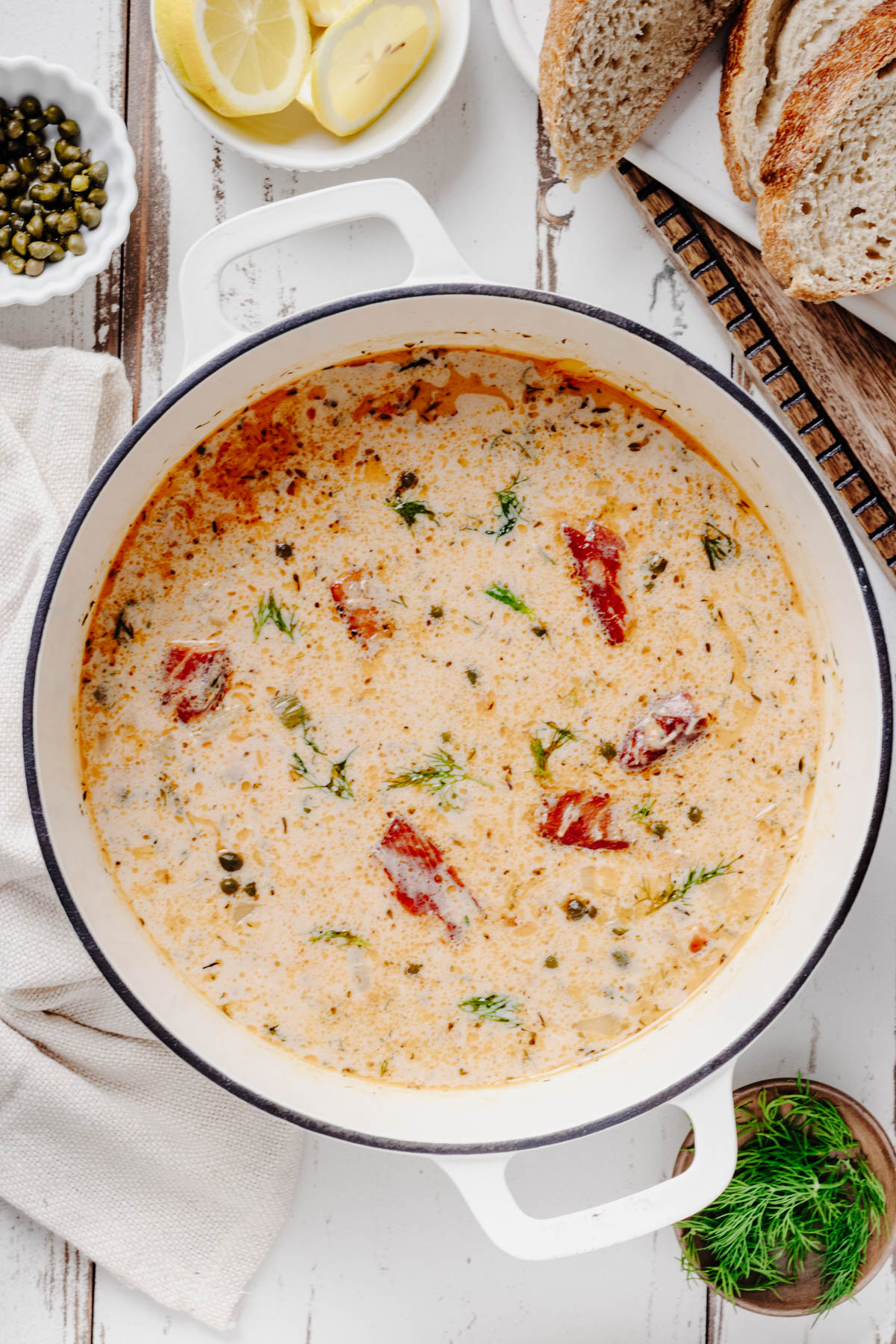 Creamy smoked salmon chowder with potatoes, capers, and fresh dill in a white Dutch oven, surrounded by lemon and crusty bread.