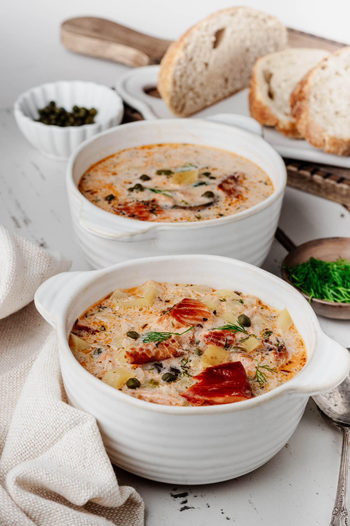 Two bowls of creamy smoked salmon chowder with potatoes, capers, and fresh dill, served with crusty bread.