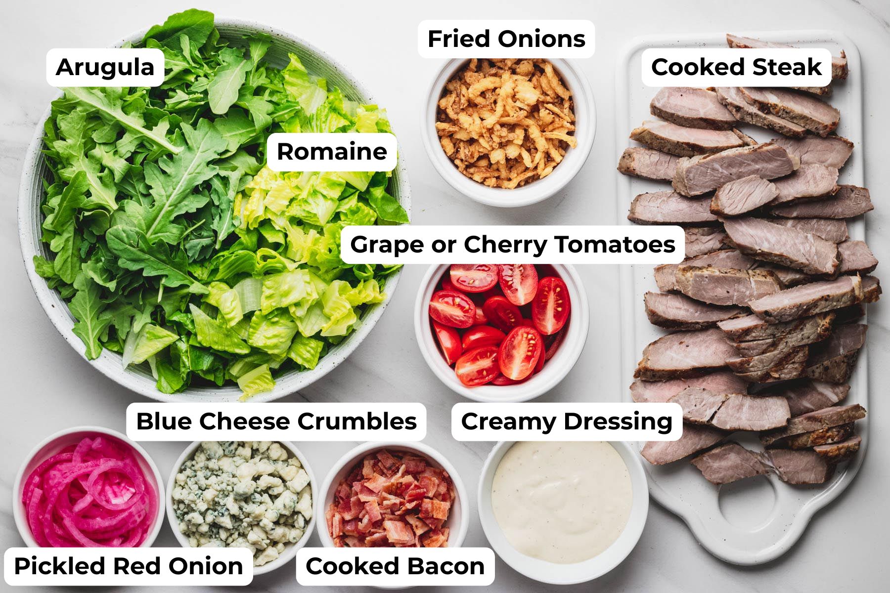 Ingredients for steakhouse salad arranged on a countertop, including romaine, arugula, cooked sirloin steak, bacon, blue cheese crumbles, pickled red onions, grape tomatoes, crispy fried onions, and creamy steakhouse dressing. Process photo for a homemade Steakhouse Salad recipe from Heavenly Home Cooking.