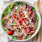An overhead view of a steakhouse salad made with crisp romaine, baby arugula, sliced sirloin steak, grape tomatoes, quick pickled red onions, blue cheese crumbles, crispy bacon, crispy fried onions, and creamy steakhouse dressing, served in a large shallow bowl with wooden salad servers.
