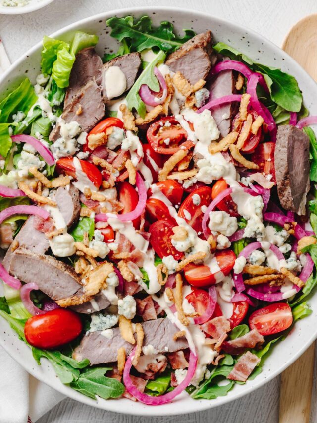 An overhead view of a steakhouse salad made with crisp romaine, baby arugula, sliced sirloin steak, grape tomatoes, quick pickled red onions, blue cheese crumbles, crispy bacon, crispy fried onions, and creamy steakhouse dressing, served in a large shallow bowl with wooden salad servers.