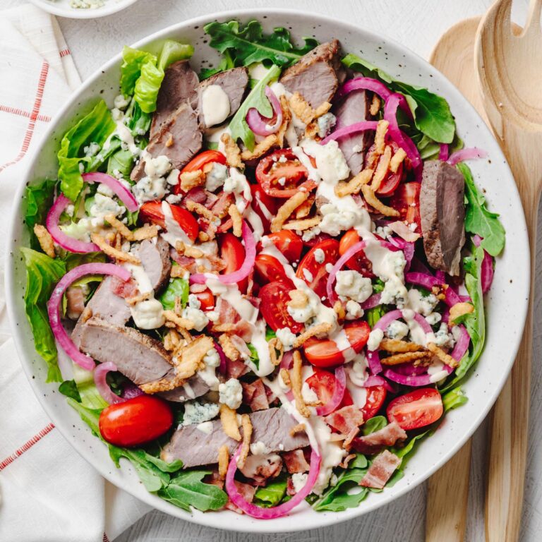 An overhead view of a steakhouse salad made with crisp romaine, baby arugula, sliced sirloin steak, grape tomatoes, quick pickled red onions, blue cheese crumbles, crispy bacon, crispy fried onions, and creamy steakhouse dressing, served in a large shallow bowl with wooden salad servers.