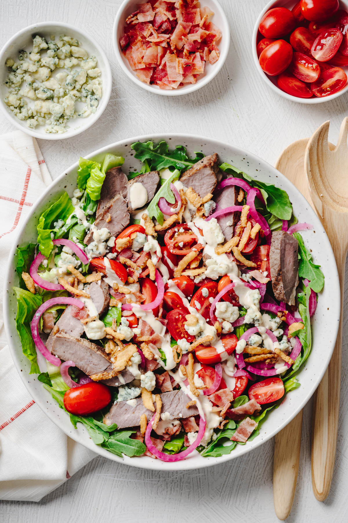 A steakhouse salad topped with sliced sirloin steak, grape tomatoes, quick pickled red onions, blue cheese crumbles, crispy bacon, crispy fried onions, and creamy steakhouse dressing, served in a large bowl with bowls of extra toppings nearby.