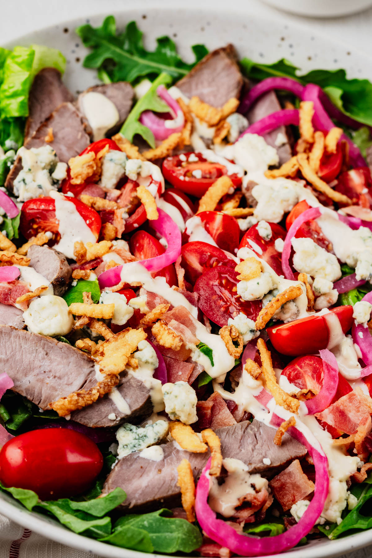 Close-up of a steakhouse salad with sliced sirloin steak, crisp romaine, baby arugula, grape tomatoes, quick pickled red onions, blue cheese crumbles, crispy bacon, crispy fried onions, and creamy steakhouse dressing.
