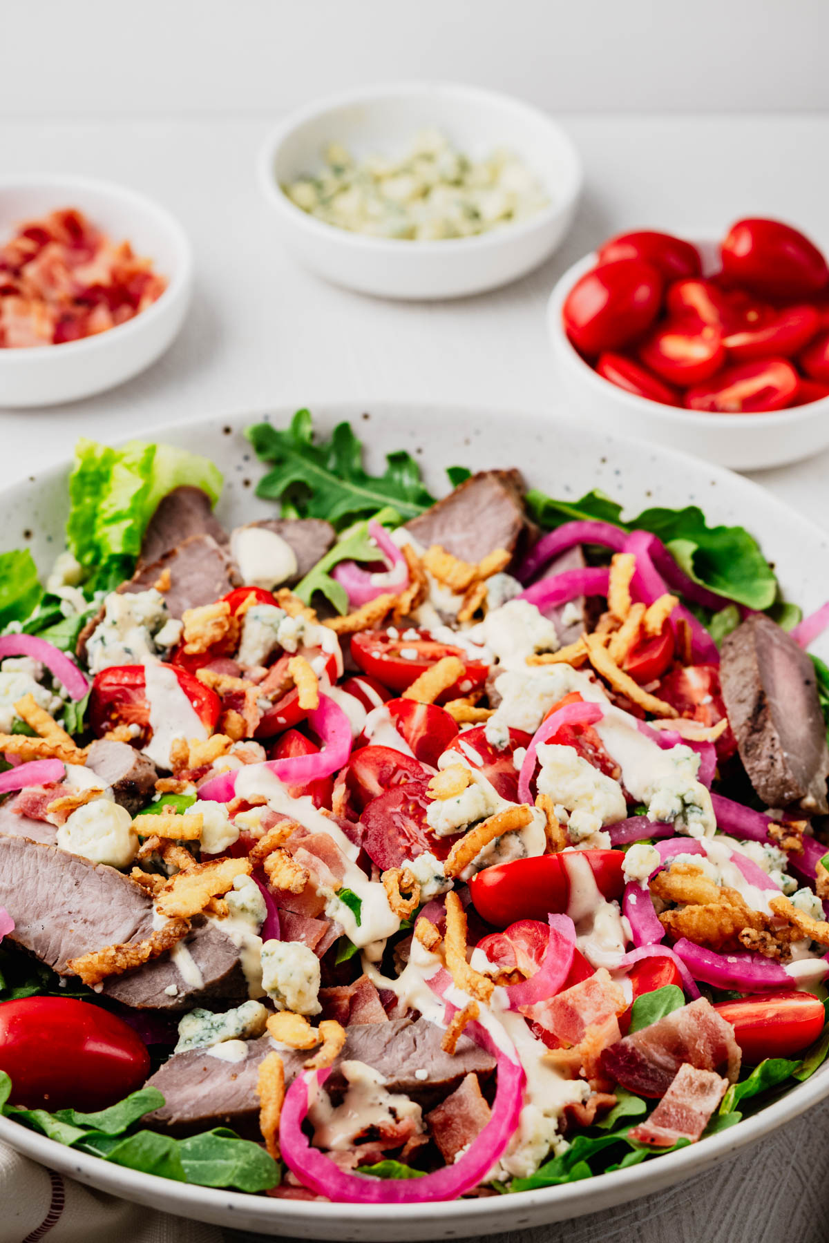 A large bowl of steakhouse salad with sliced sirloin steak, crisp romaine, baby arugula, grape tomatoes, quick pickled red onions, blue cheese crumbles, crispy bacon, crispy fried onions, and creamy steakhouse dressing, served with wooden salad servers.