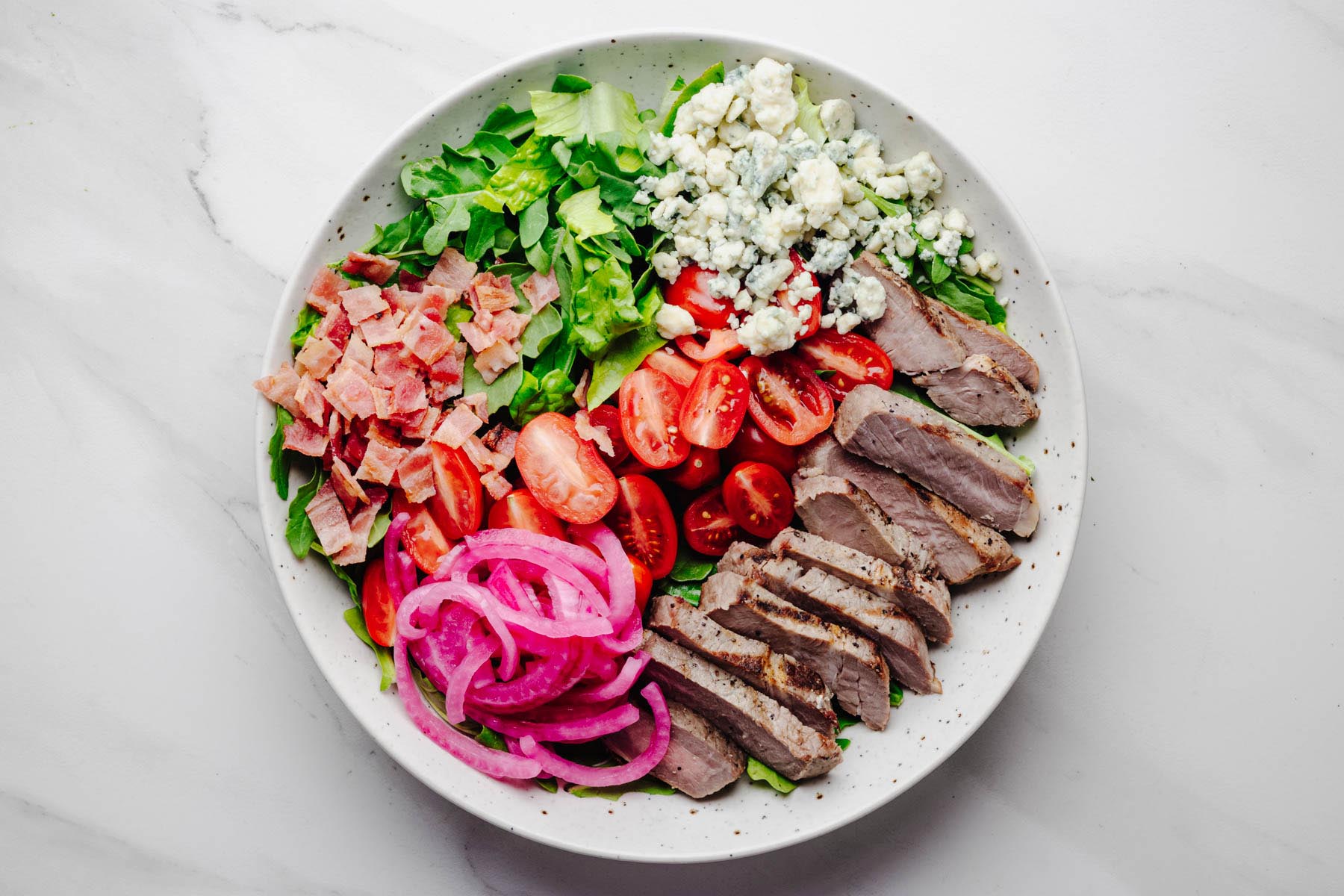 Steakhouse salad assembled with romaine, arugula, sliced sirloin steak, tomatoes, bacon, blue cheese, and pickled red onions before adding the dressing. Process photo for a homemade Steakhouse Salad recipe from Heavenly Home Cooking.