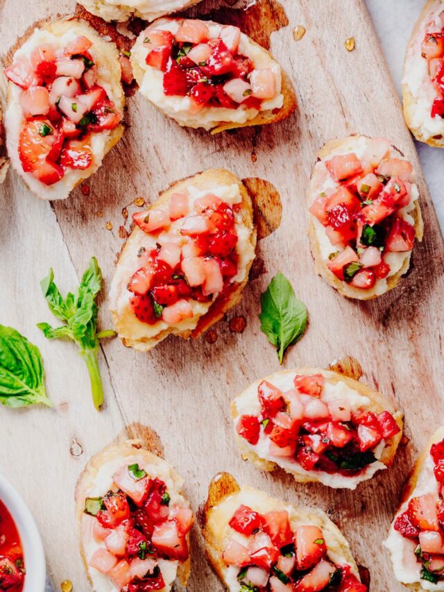 Overhead view of strawberry bruschetta made with toasted baguette slices topped with whipped ricotta and a fresh strawberry basil mixture, arranged on a rustic wooden board with fresh basil leaves and a bowl of strawberry topping. Featured image for a homemade strawberry bruschetta recipe from Heavenly Home Cooking.