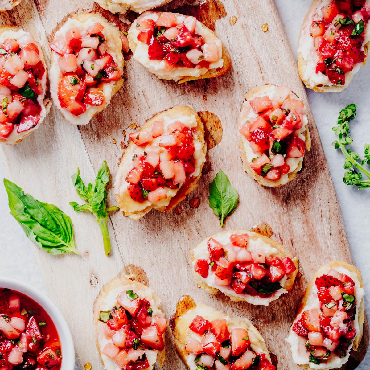 Overhead view of strawberry bruschetta made with toasted baguette slices topped with whipped ricotta and a fresh strawberry basil mixture, arranged on a rustic wooden board with fresh basil leaves and a bowl of strawberry topping. Featured image for a homemade strawberry bruschetta recipe from Heavenly Home Cooking.