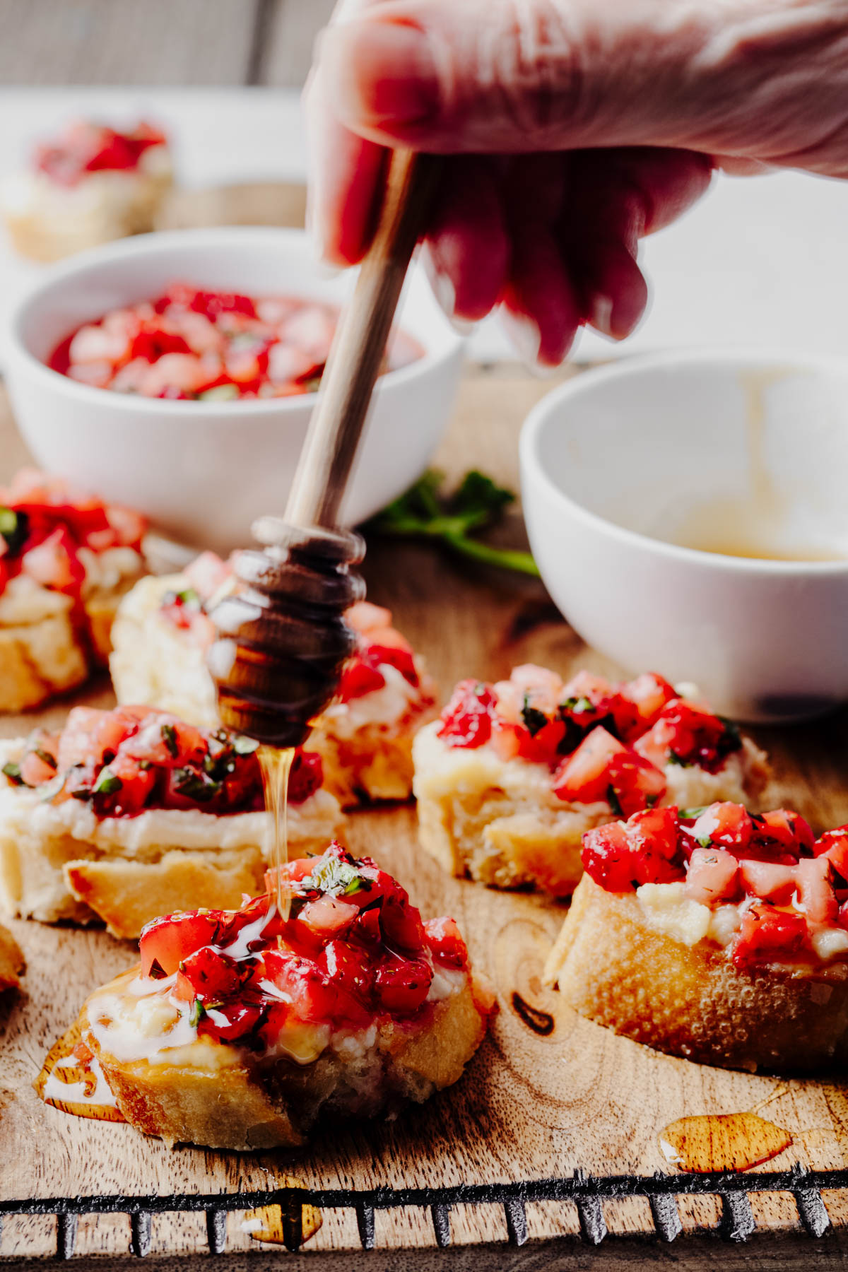 Close-up of strawberry bruschetta topped with whipped ricotta as honey is drizzled over the toasted baguette slices from a wooden honey dipper.