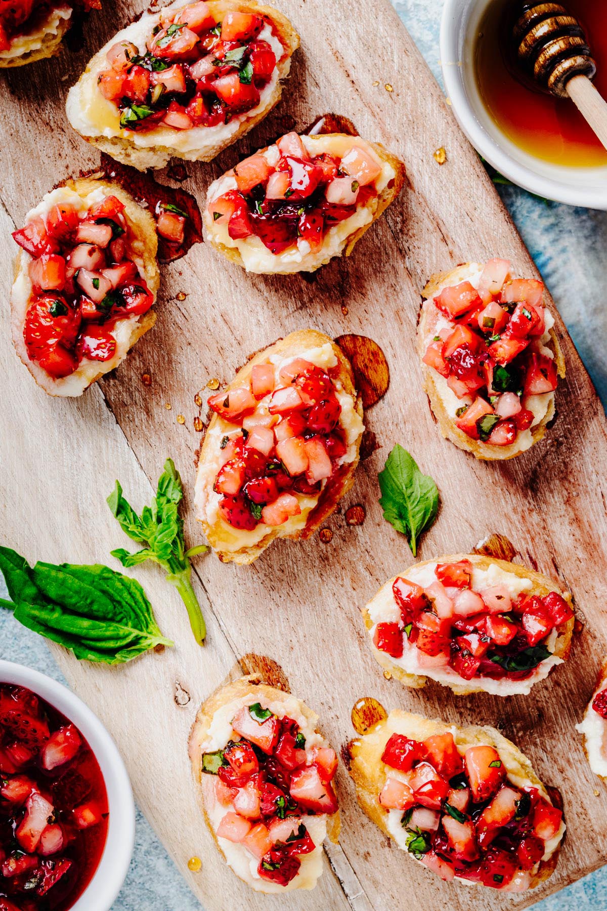 Overhead view of strawberry bruschetta with whipped ricotta on toasted baguette slices arranged on a wooden serving board with fresh basil, a bowl of honey, and a bowl of strawberry topping.