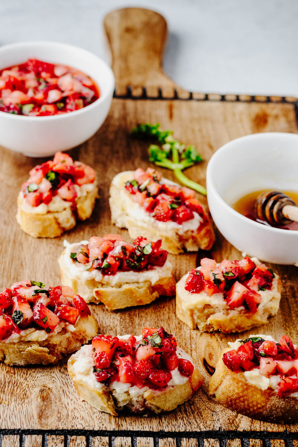 Strawberry bruschetta with whipped ricotta served on toasted baguette slices on a wooden serving board with bowls of honey and strawberry topping in the background.
