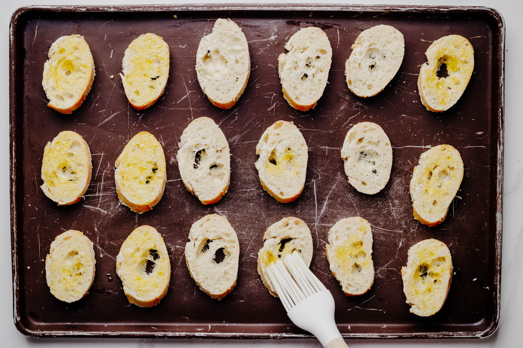Olive oil being brushed onto one side of sliced baguette rounds arranged on a baking sheet before toasting.