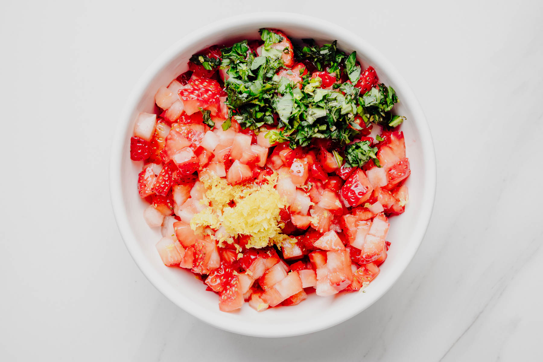 Diced strawberries, chopped fresh basil, lemon zest, honey, and lemon juice in a white bowl before being mixed together.