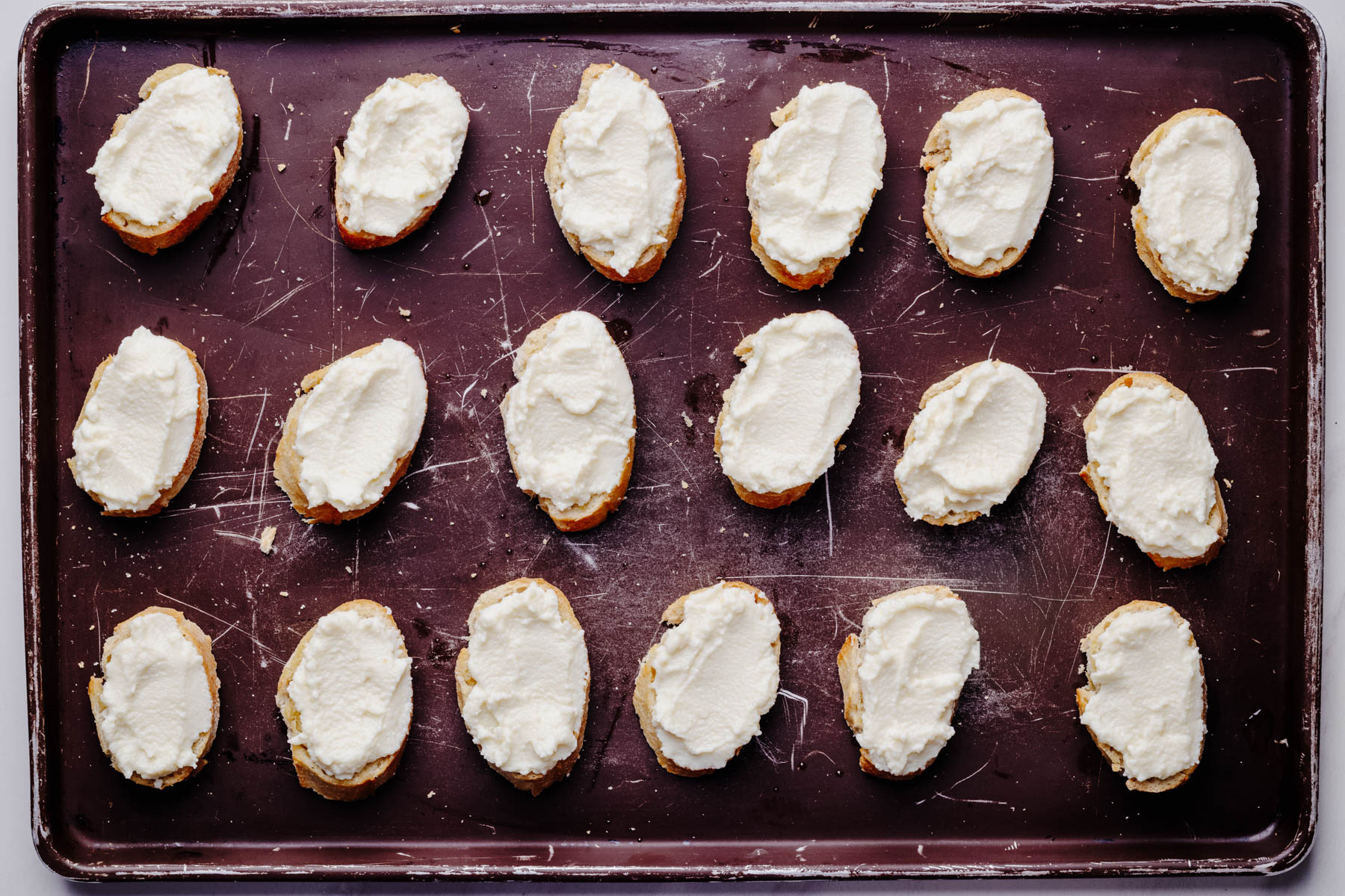 Toasted baguette slices spread with whipped ricotta and arranged on a baking sheet.