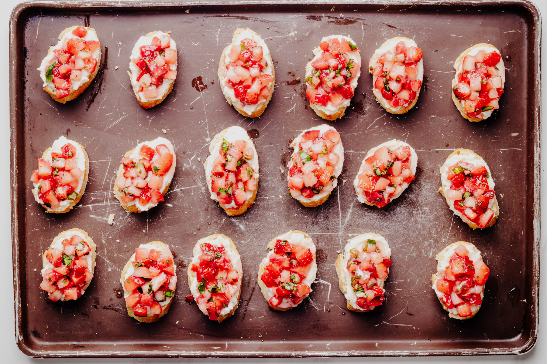 Toasted baguette slices topped with whipped ricotta and a fresh strawberry basil mixture on a baking sheet.