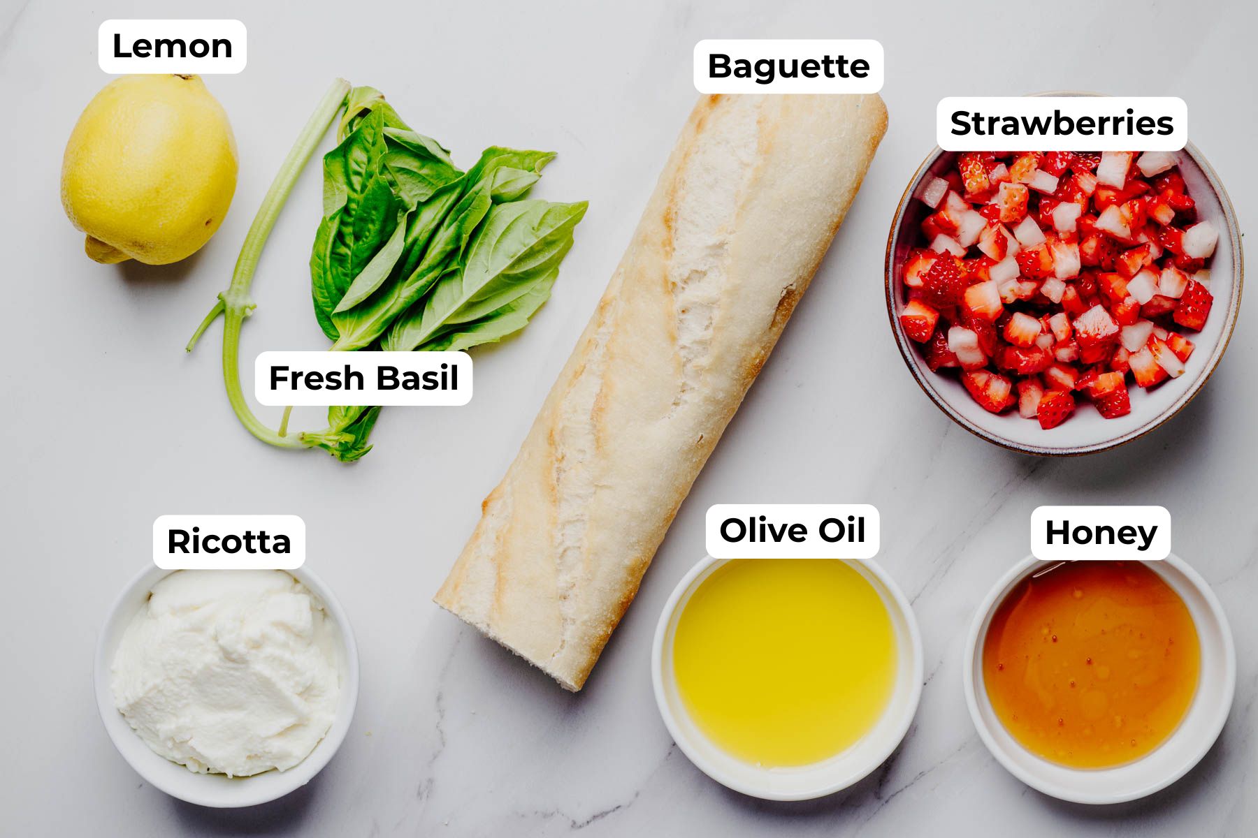 Labeled ingredient photo for strawberry bruschetta showing a baguette, ricotta cheese, diced strawberries, fresh basil, a lemon, olive oil, and honey arranged on a white surface.