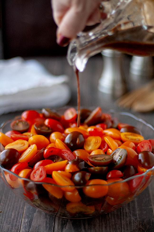 A hand pours a dark balsamic vinaigrette over a bowl of colorful cherry tomatoes in shades of red, orange, and purple, capturing the start of a fresh tomato salad.