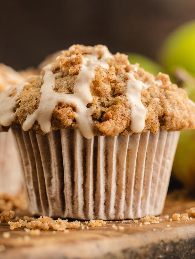 Apple streusel muffin with cinnamon crumb topping and maple icing, with apples softly blurred in the background.