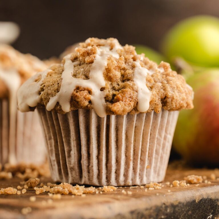 Apple streusel muffin with cinnamon crumb topping and maple icing, with apples softly blurred in the background.