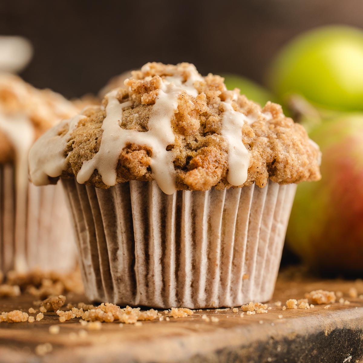 Apple streusel muffin with cinnamon crumb topping and maple icing, with apples softly blurred in the background.