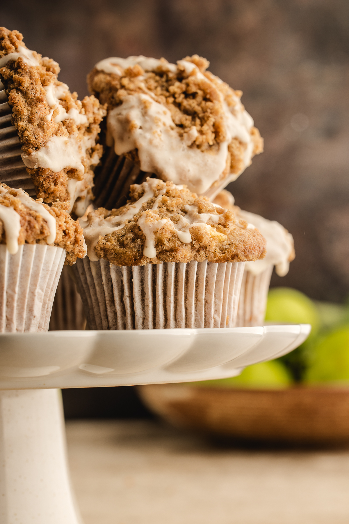 Several apple streusel muffins with maple icing arranged on a white cake stand with apples in the background.