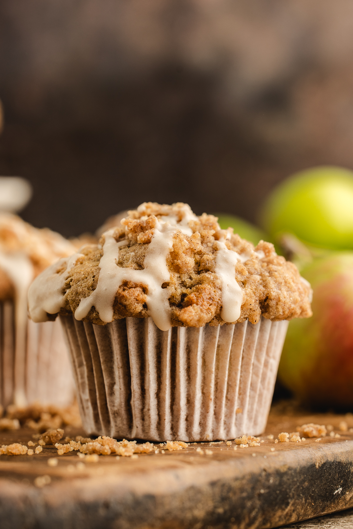 Apple streusel muffin with a tall crumb topping and maple icing, with apples softly blurred in the background.