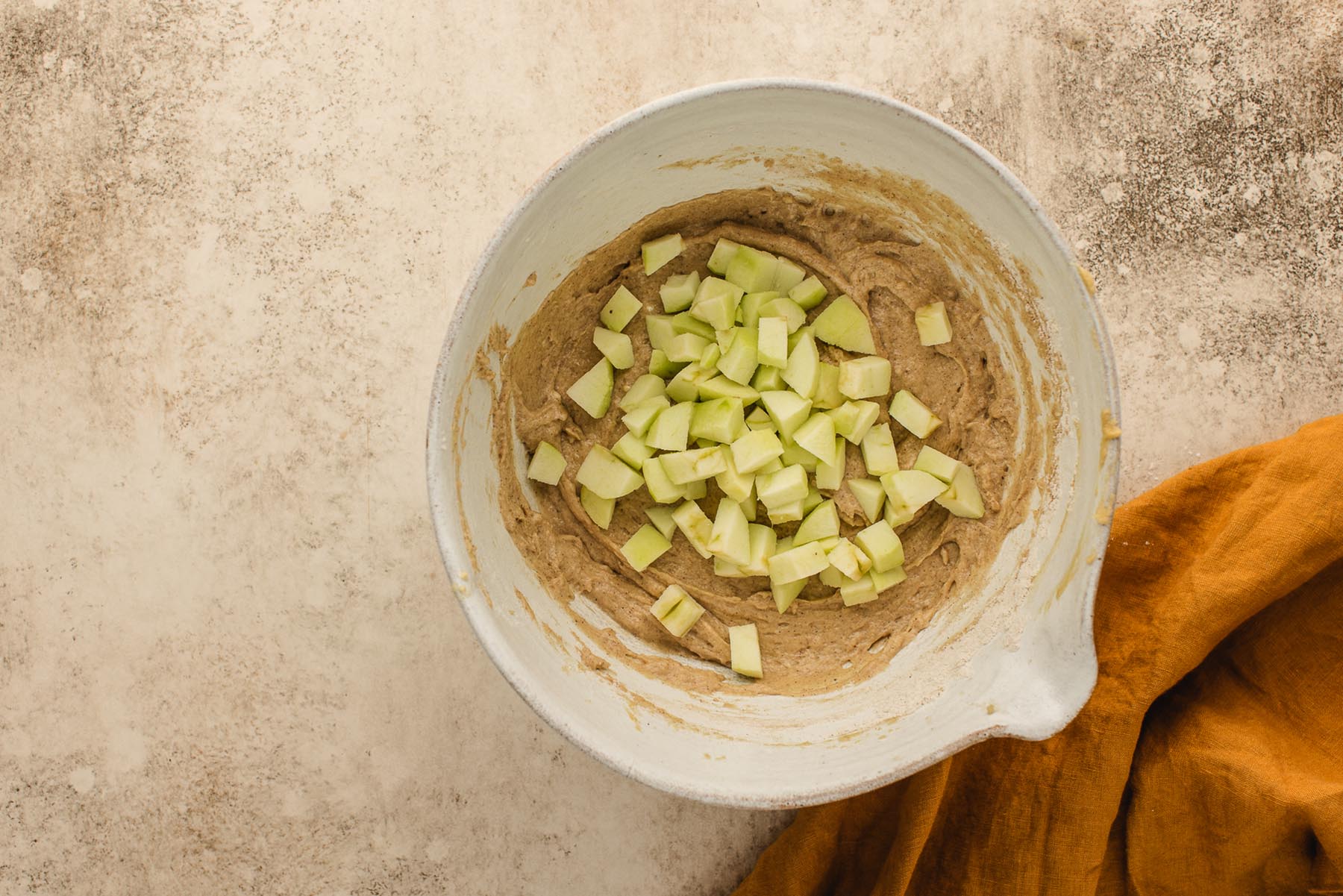 Diced green apples added to thick cinnamon muffin batter in a white mixing bowl.