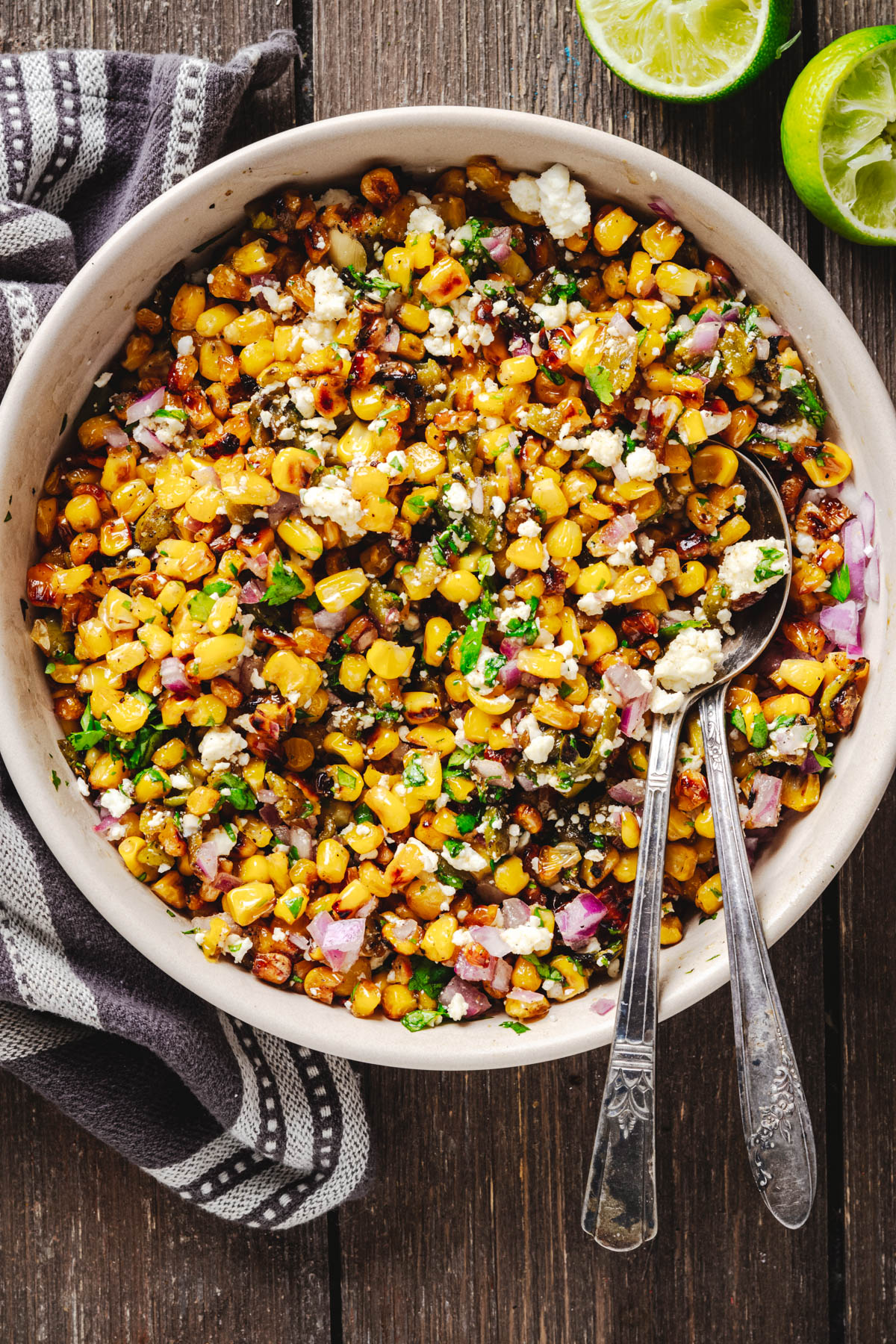 Bowl of Hatch chile corn salad with charred corn, roasted green chiles, Cotija, cilantro, and red onion, served with lime and spoons.