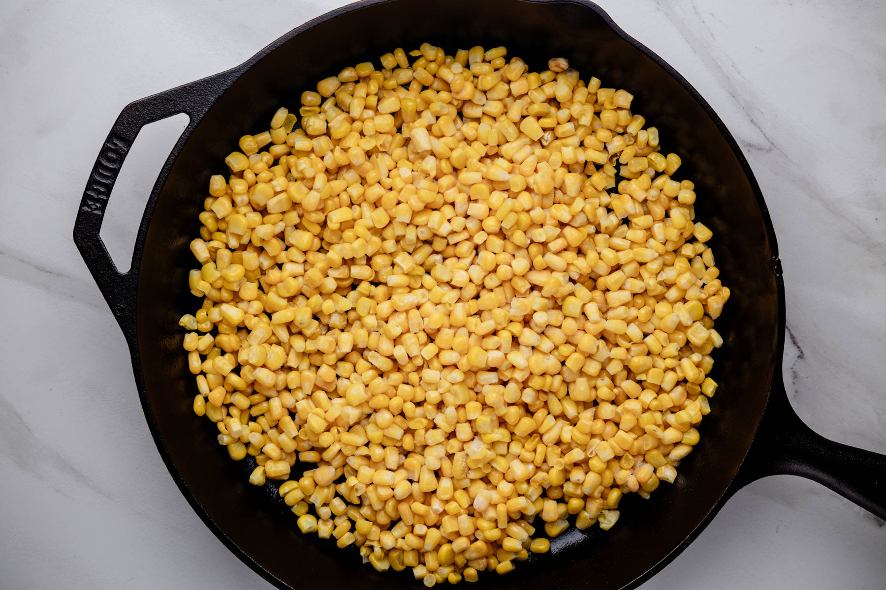 Thawed corn kernels spread in an even layer in a large cast-iron skillet.
