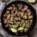 Overhead view of Hatch chile steak bites garnished with cilantro and lime wedges in a cast-iron skillet.